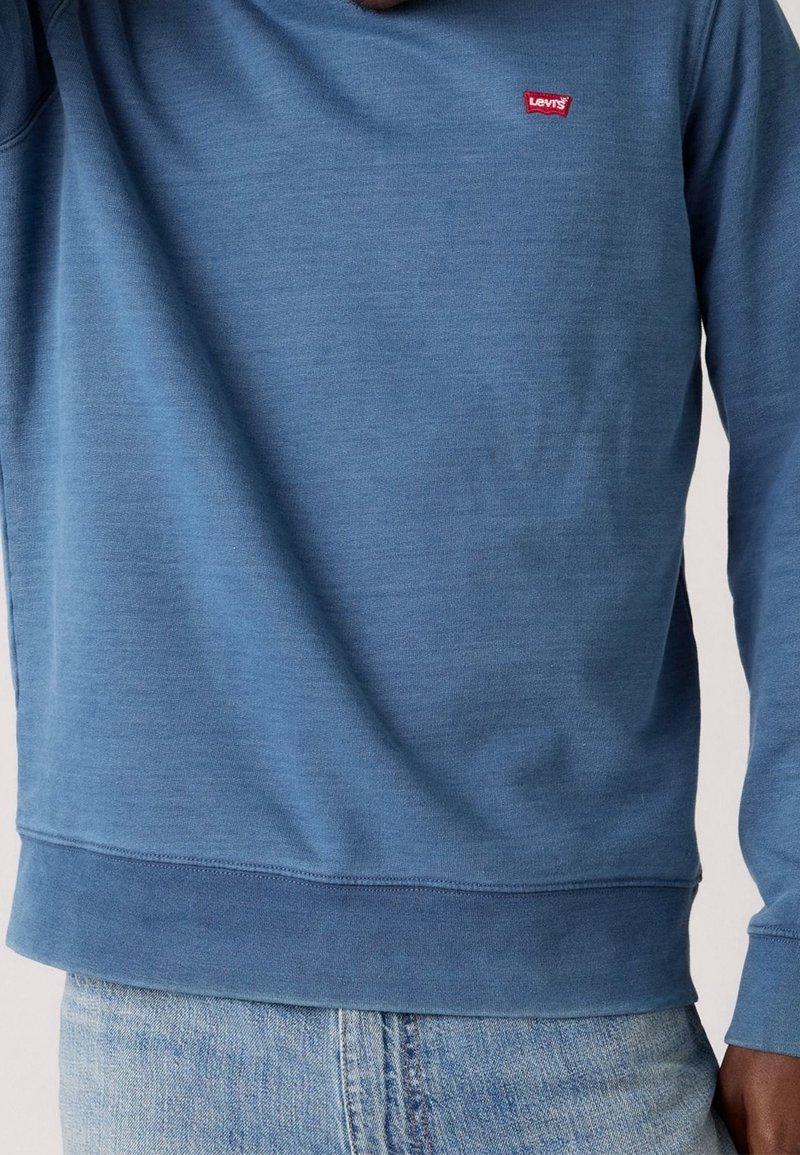 Homme portant un sweat-shirt bleu Levi's et un jean bleu clair, montrant le torse avant et la zone du bras.