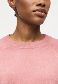 Femme portant un pull ras du cou en tricot rose clair, visible des épaules aux lèvres sur un fond clair uni.