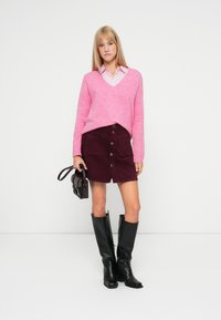 Rózsaszín V nyakú pulóver, bordó kordbársony gombos szoknya, fekete térdigt érő csizmák és egy kis fekete kézitáska. Sima textúrák és testhezálló fazon.