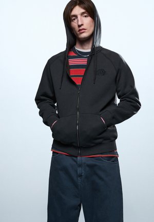 Mladi človek, ki nosi črno jopa z zadrgo čez rdeče-črni črtasti pulover, temne široke hlače, roke v žepih jopice, nevtralen izraz na obrazu.