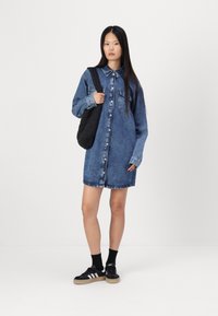 Vestito camicia in denim di un blu medio con botttoni frontali, maniche lunghe, due tasche sul petto e orlo sfilacciato. Abbinato a sneakers nere e una borsa trapuntata.