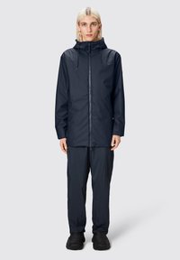 Giacca impermeabile navy con zipper frontale, cappuccio regolabile e maniche lunghe, abbinata a pantaloni coordinati e scarpe scure. Texture liscia.