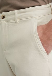 Lichtbeige katoenen broeken met een ritssluiting, enkele knoopsluiting en zijzakken. De stof heeft een gladde textuur en een subtiele diagonale weving.