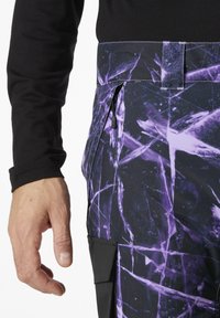 Zwarte langemouwen shirt gecombineerd met paarse en zwarte met patronen broek met een zijzak en ritssluiting, die hoekige, abstracte ontwerpen benadrukt.
