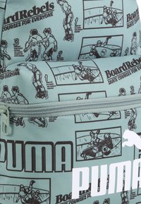 Zaino in tessuto verde con grafiche nere che raffigurano figure di skateboard, dotato di un compartimento con cerniera e logo Puma bianco.