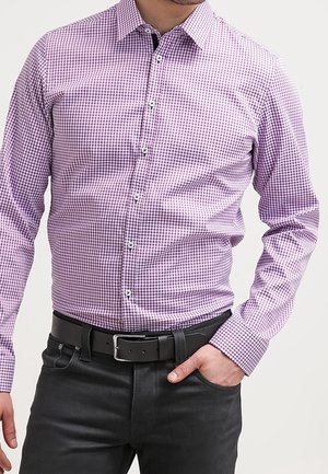 Uomo che indossa una camicia aderente a quadri viola e bianchi infilata in pantaloni neri con cintura nera, mano in tasca.