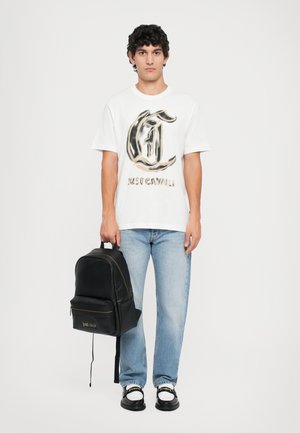 Hvit T-skjorte med kort erme som har et stort svart og gull logo trykk, paret med lyseblå jeans og en svart ryggsekk. Svarte sko er brukt.