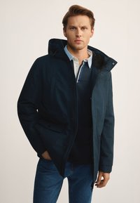 Granatowa parka z kapturem, zapinana na zamek, z wieloma kieszeniami i miękką podszewką. Nosi się ją na jasnoniebieską koszulę z kołnierzykiem.