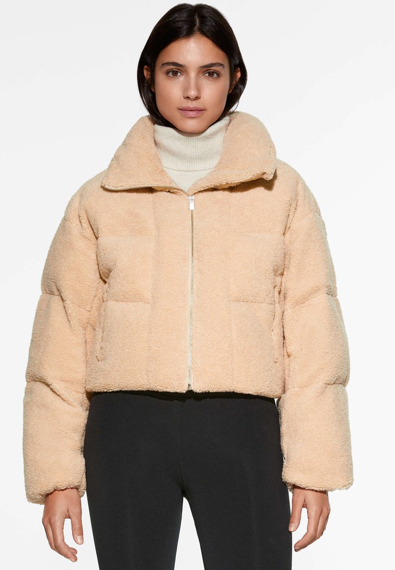 Pfirsichfarbene, kurz geschnittene Puffermütze aus strukturiertem Gewebe mit hohem Kragen und durchgehendem Reißverschluss. Sie verfügt über große gesteppte Einsätze und Seitentaschen.