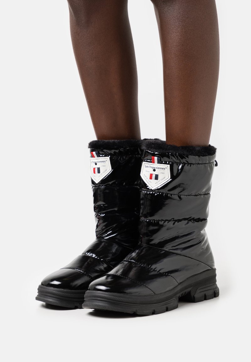 Bottes noires brillantes en style doudoune avec une bordure en fausse fourrure, des semelles en caoutchouc et des détails de logo en forme de bouclier, présentant un design matelassé.