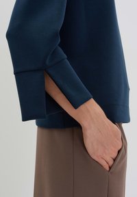 Dark blue sweatshirt with a crew neck, featuring slit cuffs and smooth texture. Brown trousers with side pockets visible, creating a layered look.