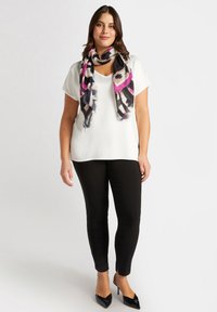 Top blanco de manga corta combinado con pantalones negros ajustados y zapatos de tacón negros. Una bufanda colorida con estampado añade contraste alrededor del cuello.