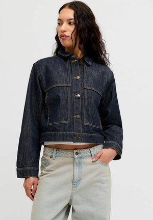 Femme aux longs cheveux bruns portant une veste en jean courte bleu foncé et un jean taille haute bleu clair, debout avec une main dans la poche.