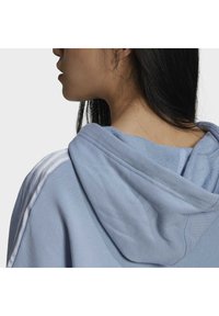 Kvinna med långt svart hår som bär en ljusblå huvtröja med vita ränder på axeln, visad från sidan och bakifrån.