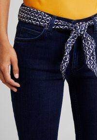Los jeans de mezclilla de un azul oscuro presentan un cinturón de patrón en azul marino y blanco atado en un lazo. La tela tiene una textura suave con un clásico diseño de cinco bolsillos.