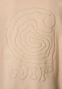 Fleece audums gaiši bēšā krāsā ar teksturētu spirāles rakstu gaiši zaļā diegā, blakus ir vārds "WIP" biezā, neregulārā fontā.