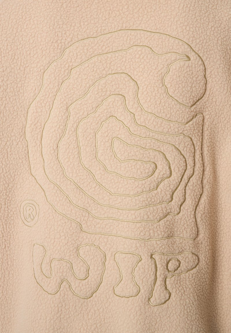 Fleece-tyg i ljusbeige med ett strukturerat, spiralformat mönster i ljusgrön tråd, tillsammans med ordet "WIP" i en tjock, oregelbunden typografi.