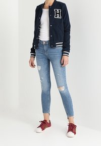 Námornická bombarďácká bunda s bílými pruhy a vyšitým "H", bílým tričkem, distressed sv ětle modrými skinny džíny a bordo teniskami.