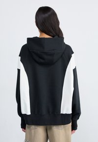 H hoodie noir avec des panneaux latéraux blancs, doté d'une capuche avec cordon de serrage et de poignets côtelés. Tissu doux et coupe décontractée, conçu pour le confort.