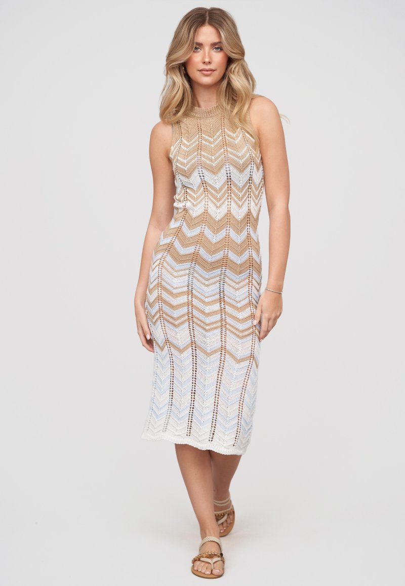 Jeune femme aux longs cheveux blonds portant une robe tricotée sans manches beige et blanche à motif chevrons et des sandales, debout devant un fond uni.