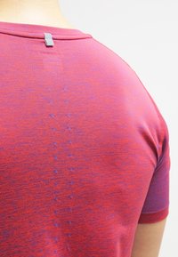 Camiseta deportiva de manga corta en punto jaspeado rojo y púrpura, con un reverso liso que presenta patrones de puntos en contraste y un pequeño lazo en el escote.