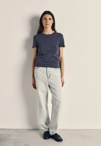 Woman standing against light wall wearing navy blue t-shirt, light blue jeans, and black loafers, with a neutral expression.