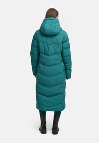 Dlhý tyrkysový prešívaný kabát s kapucňou, ktorý má prešívaný vzor a mierne sploštený pás, skombinovaný s čiernymi členkovými čižmami.