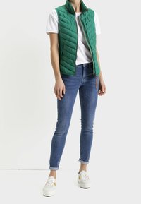 Veste matelassée verte à col montant, portée sur un t-shirt blanc, associée à un jean skinny bleu et des baskets blanches.