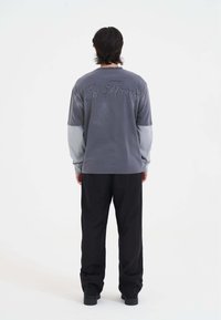 Langarmshirt van grijze stof met lichtergrijze mouwen, met geborduurd logo op de achterkant. Gekocht met zwarte broek en schoenen.