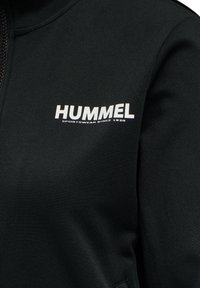 Czarna sportowa kurtka z zamkiem błyskawicznym, miękką teksturą i wyraźnym białym logo "HUMMEL" z napisem "SPORTSWEAR SINCE 1920" pod spodem.