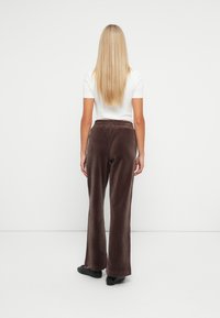 Brūnas, platās sporta bikses mīkstā velūra audumā ar elastīgu jostasvietu, kombinētas ar baltu īsām piedurknēm topu un melnām kurpēm.