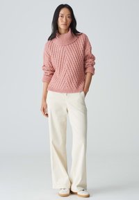 Rosa gestrickter Pullover mit hohem Kragen und strukturiertem Muster, kombiniert mit weiten weißen Cordhosen und weißen Schuhen mit braunen Akzenten.