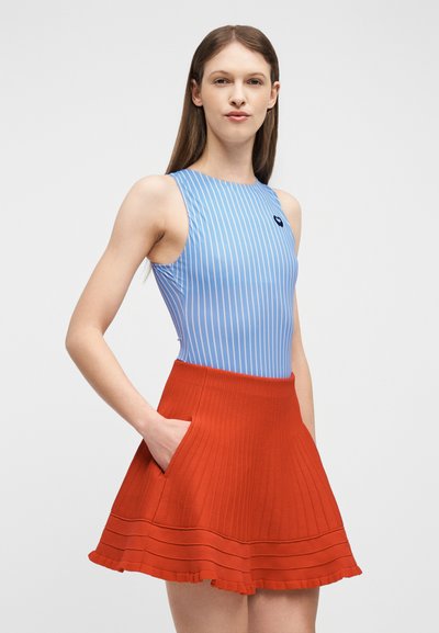 Femme aux longs cheveux bruns portant un haut sans manches rayé bleu et blanc et une jupe évasée rouge avec des poches, se tenant devant un fond uni.