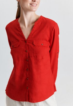 Kobieta ubrana w czerwoną bluzkę na guziki z długim rękawem, z dwoma kieszeniami na piersi i dekoltem w kształcie litery V, stojąca na tle jednolitego jasnego tła.