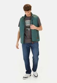 Polstret grøn vest med høj krave, lynlås og sidelommer, lagdelt over en grå grafik t-shirt og parret med blå jeans.