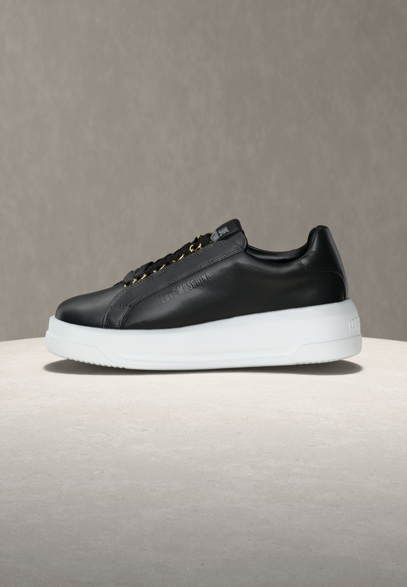 Μαύρο δερμάτινο αθλητικό παπούτσι χαμηλού τέτοιου με λευκή εξωτερική σόλα, χρυσά κρικάκια και το λογότυπο «Love Moschino» ανάγλυφο στο πλάι, τοποθετημένο σε ουδέτερη επιφάνεια.