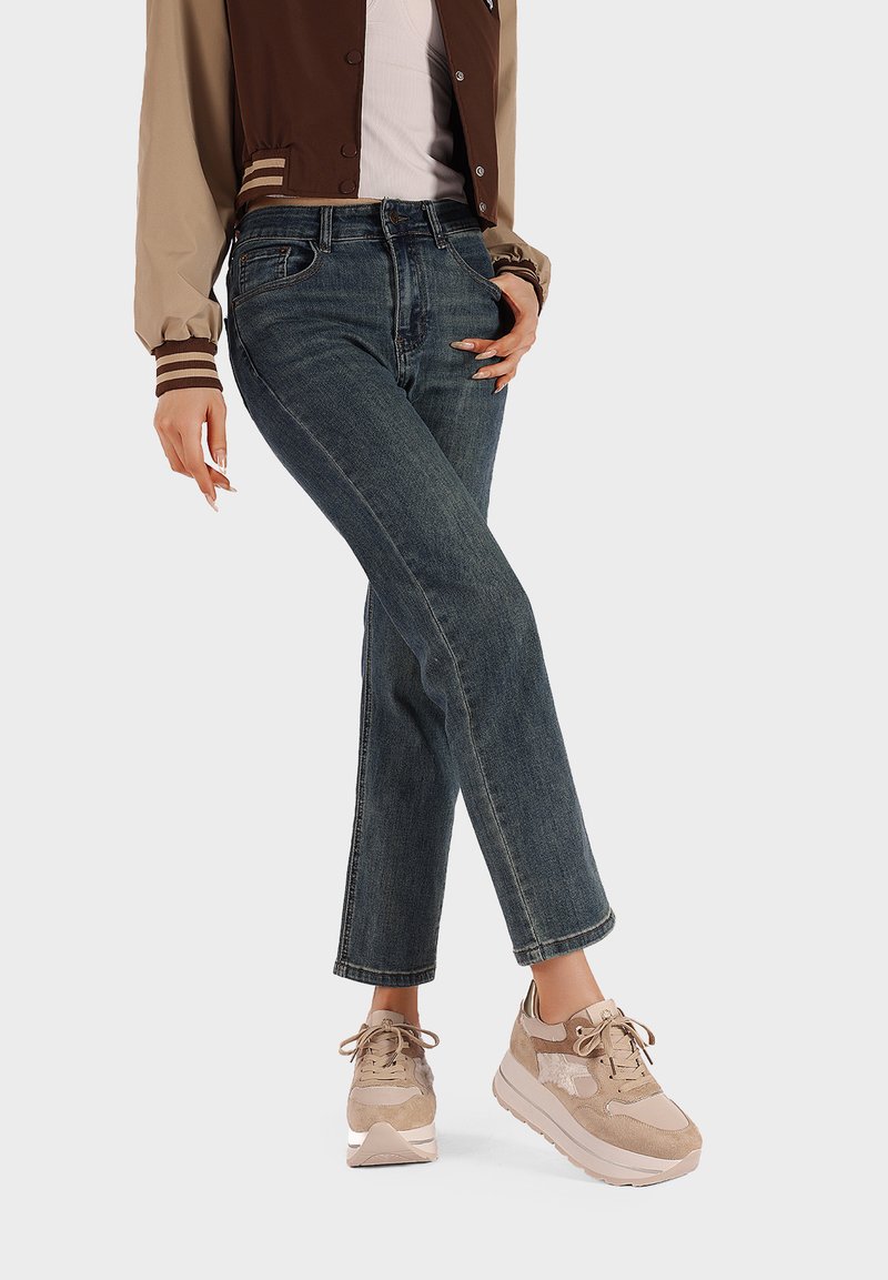 Giacca bomber beige e marrone, top bianco, jeans in denim scuri con taglio dritto e sneakers marrone chiaro con accenti testurizzati.