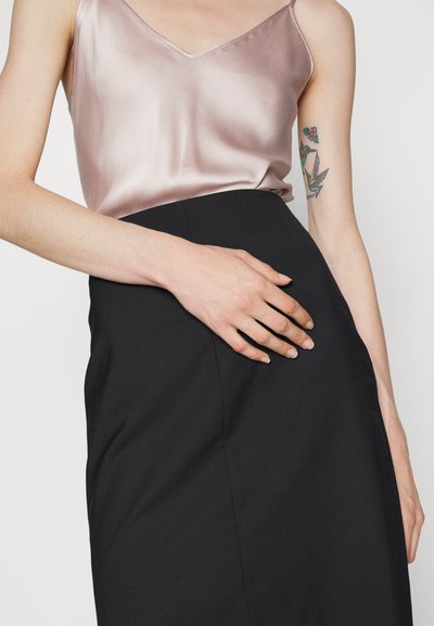 Camisole en satin rose clair avec décolleté en V, associé à une jupe noire moulante ; main reposant sur la couture latérale de la jupe. Design minimaliste.