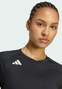 Černé sportovní tričko s kulatým výstřihem a krátkými rukávy, s bílým trojúhelníkovým logem na levém rameni. Hladká textura.
