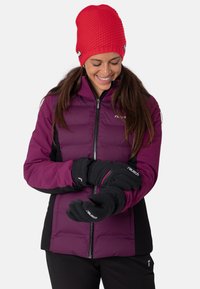 Mujer con un gorro de punto rojo, chaqueta morada y negra, ajustándose guantes negros con un detalle rosa, sonriendo y mirando hacia abajo.