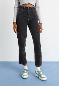 Melni augstās jostas vidus džinsi ar kontrastējošiem šuvēm, taisnu kāju dizains un pieci kabatas, apvienoti ar zaļiem un baltajiem sporta apaviem.