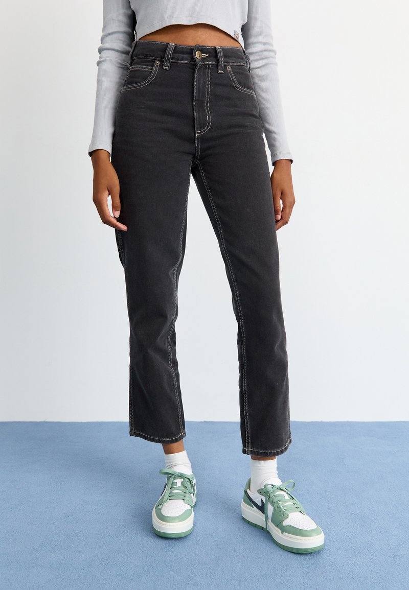 Melni augstās jostas vidus džinsi ar kontrastējošiem šuvēm, taisnu kāju dizains un pieci kabatas, apvienoti ar zaļiem un baltajiem sporta apaviem.