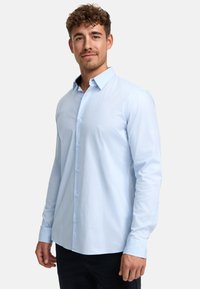 Camisa de botones de manga larga color azul claro con cuello puntiagudo, puños sencillos y tapeta frontal con botones a juego. Textura de tejido suave.