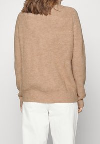 Beige gestrickter Pullover mit gerippter Textur, rundem Ausschnitt und langen Ärmeln, kombiniert mit weißen Hosen. Schlichte Rückansicht.