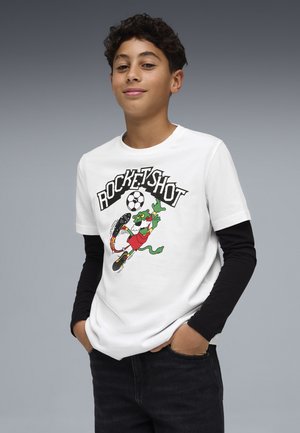 Ragazzo con capelli ricci che indossa una maglietta bianca "Rocketshot" con una lucertola dei cartoni animati che calcia un pallone da calcio infuocato, maniche lunghe nere e jeans scuri.