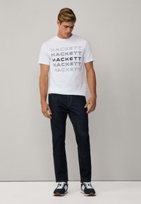 Camiseta de algodón blanca con el logotipo "HACKETT" repetido en azul marino. Combinada con unos vaqueros oscuros y zapatillas deportivas negras con detalles blancos.