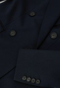 Primer plano de la manga y la parte delantera de una chaqueta azul marino oscuro con botones negros, incluyendo cuatro botones en el puño y tres en la parte delantera.