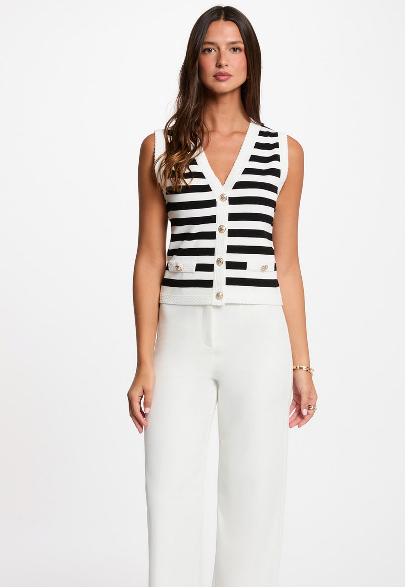 Woman wearing a sleeveless black and white horizontal striped buttoned vest with white wide-leg pants, standing against a plain background.