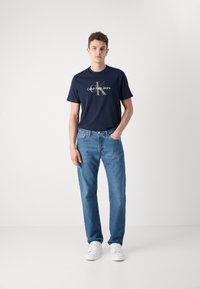 Camiseta de algodón de color navy con un logo impreso, combinada con unos jeans de denim azul claro. El modelo lleva zapatillas blancas y está de pie en un fondo neutro.