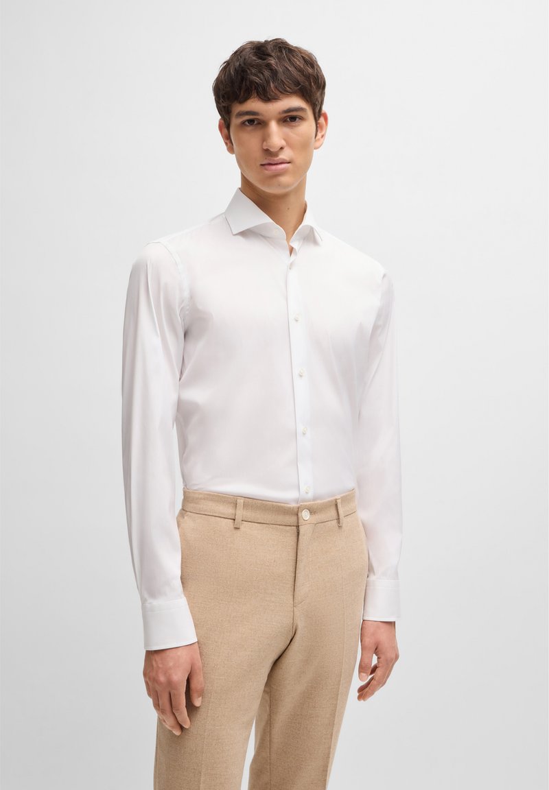 Chemise blanche à manches longues et à boutons en tissu lisse, associée à un pantalon beige clair sur mesure avec une fermeture à boutons et des poches latérales.